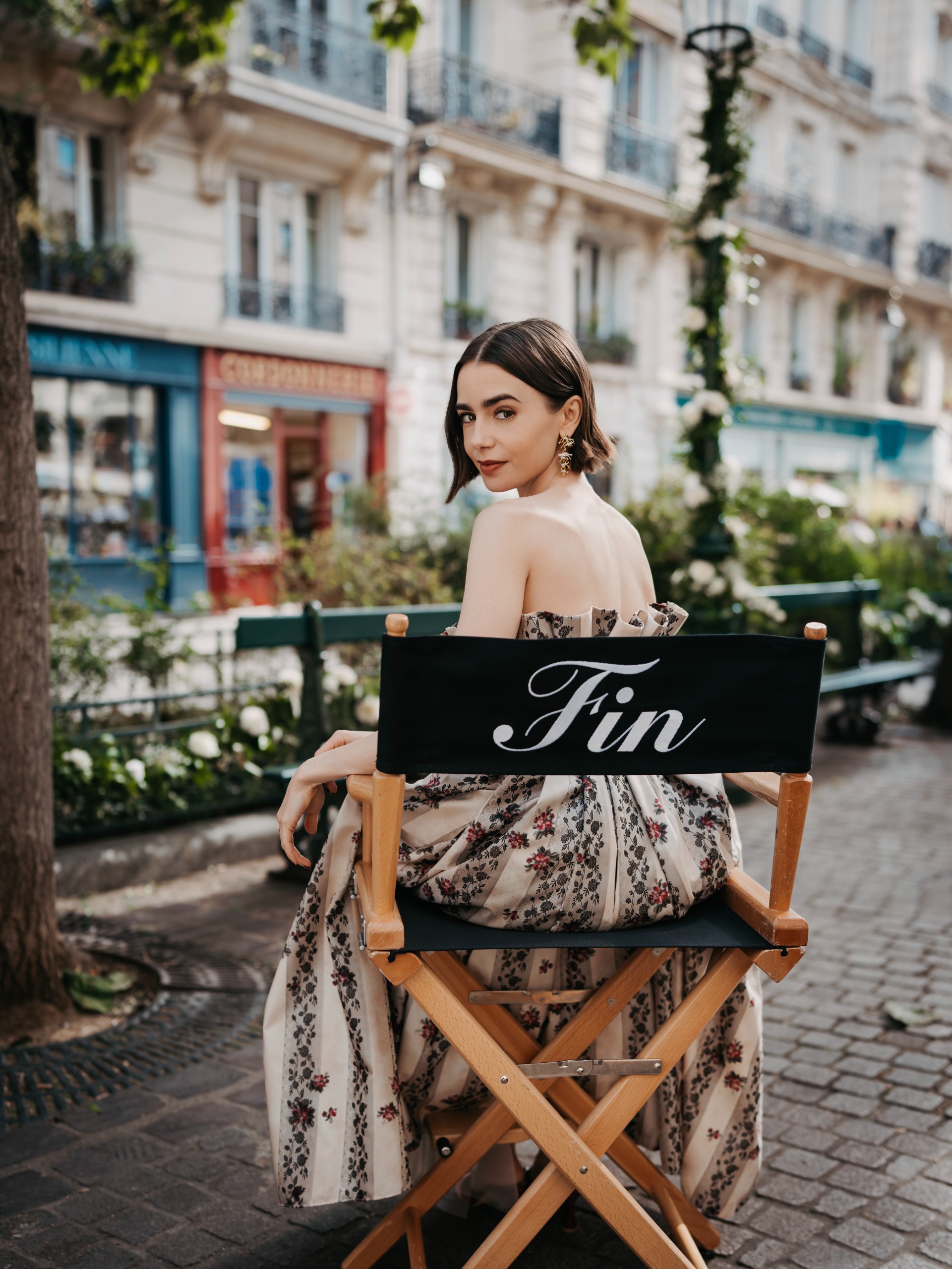 lily collins on set of the last day of emily in paris wearing her hair in a bop and a long floral dress turning over her shoulder while sitting in a director's chair that says fin