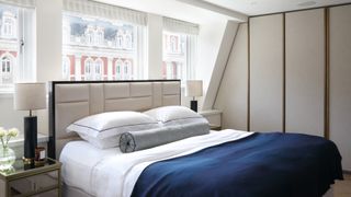 a neutral London bedroom with built-in wardrobes, a bed with a blue throw, and a bedside table, and a window view to London buildings