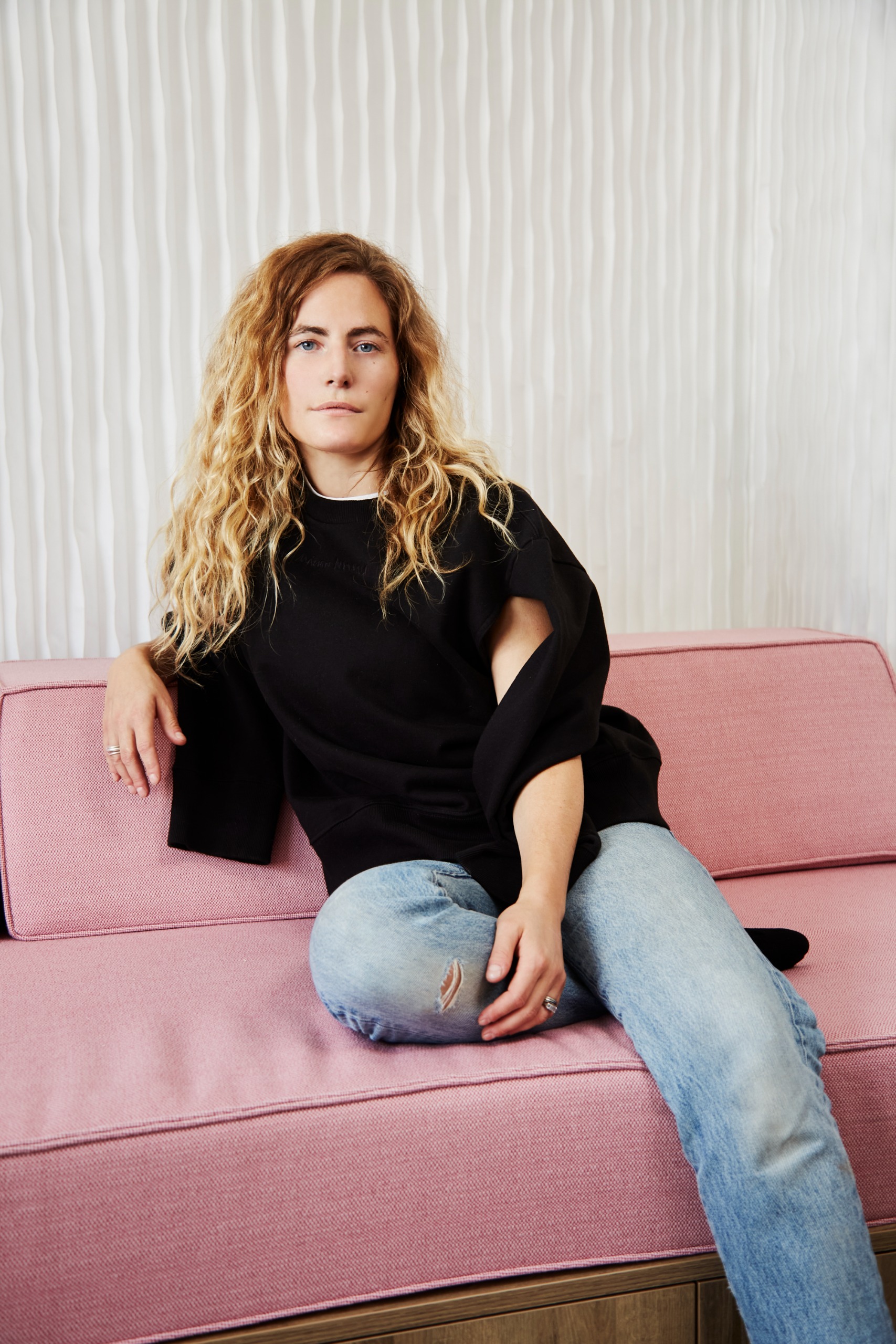 A young woman with a black top and pale denim jeans sits with her blonde curly hair free on her shoulders on a pink sofa.