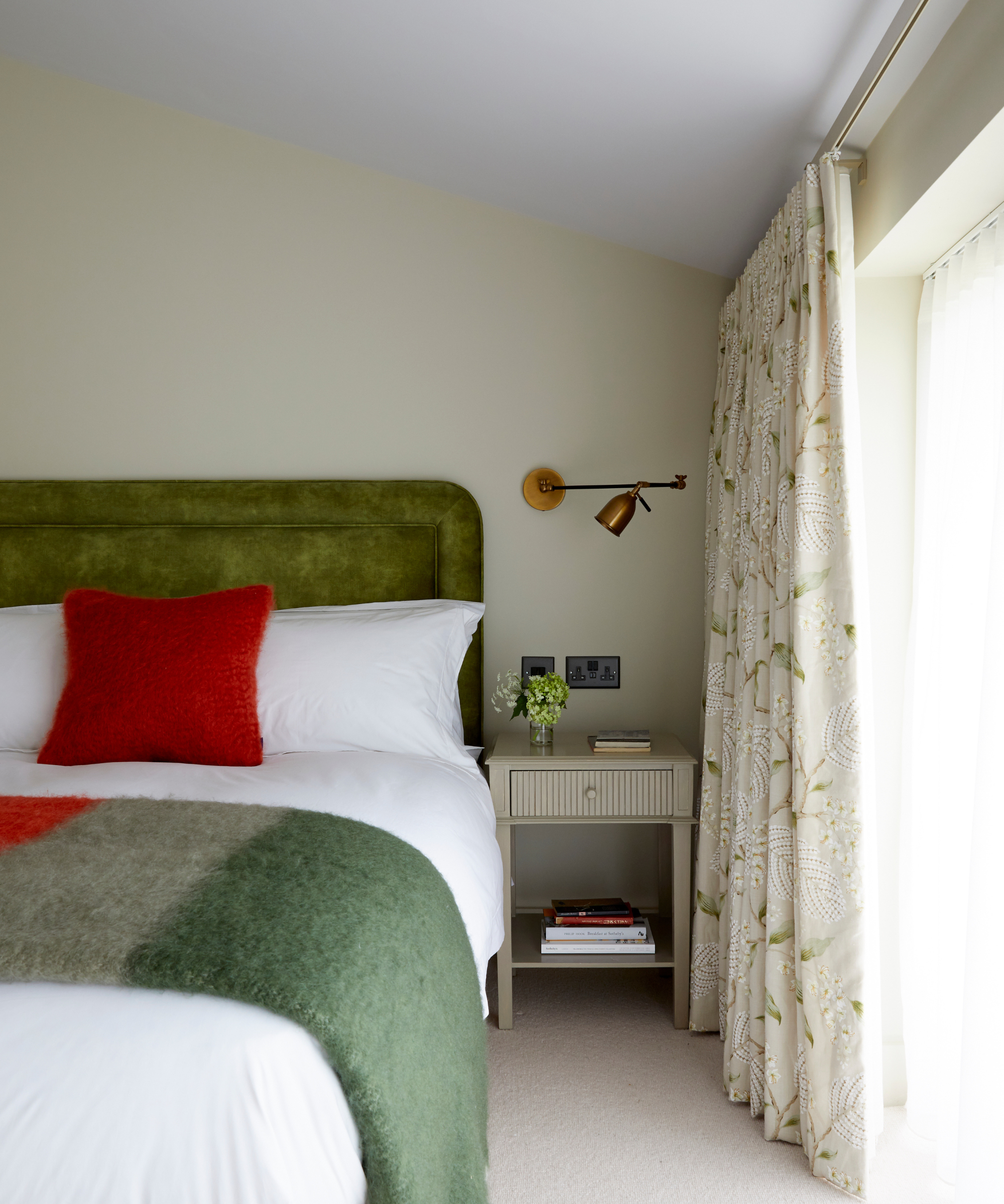 a bedroom with a green headboard, white bedding, a red pillow, a green blanket, and a brass wall sconce on the wall