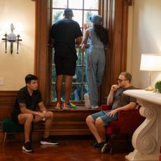 Two people stand on a windowsill and look out of a large window, as another pair sit in chairs talking, in a still from 'Got to Get Out.'