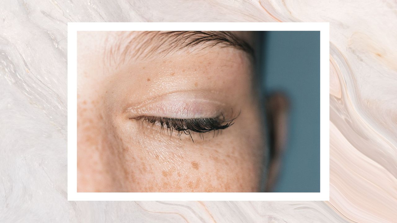 An image of a woman with freckly skin, close up to one of her eyes which is closed showing curled eyelashes on a swirly cream background