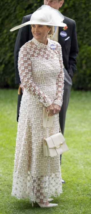 Duchess Sophie standing in grass with Prince Edward wearing a long sheer white dress and carrying a white bag at Royal Ascot