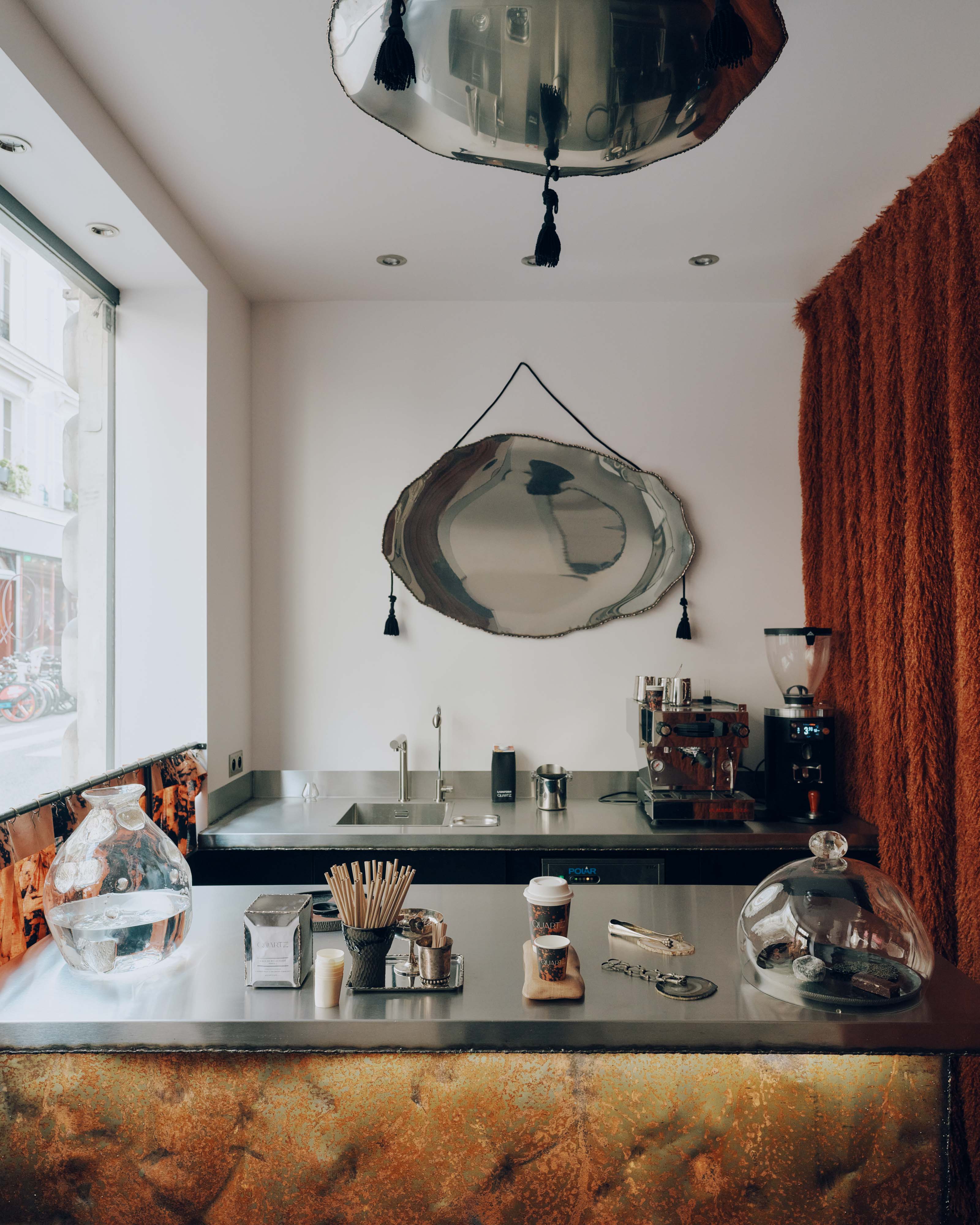 A coffee shop decorated with reflective, organically shaped chrome decor, brown curtains and wooden panels, and ceramic plates.