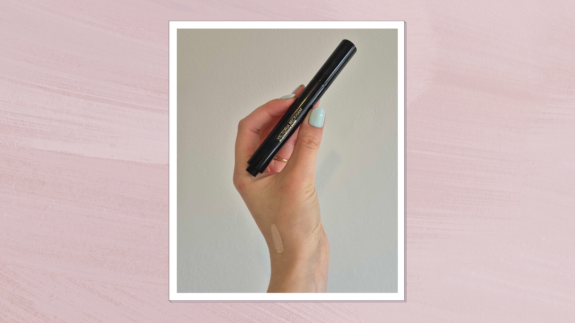 A close-up of Digital Beauty Writer, Naomi Jamieson's hand holding the The Victoria Beckham Beauty Concealer Pen with TFC8®, in front of a white wall. A swatch of the product can also be seen on the back of Naomi's hand. The image is features in pastel purple, watercolour paint-style template