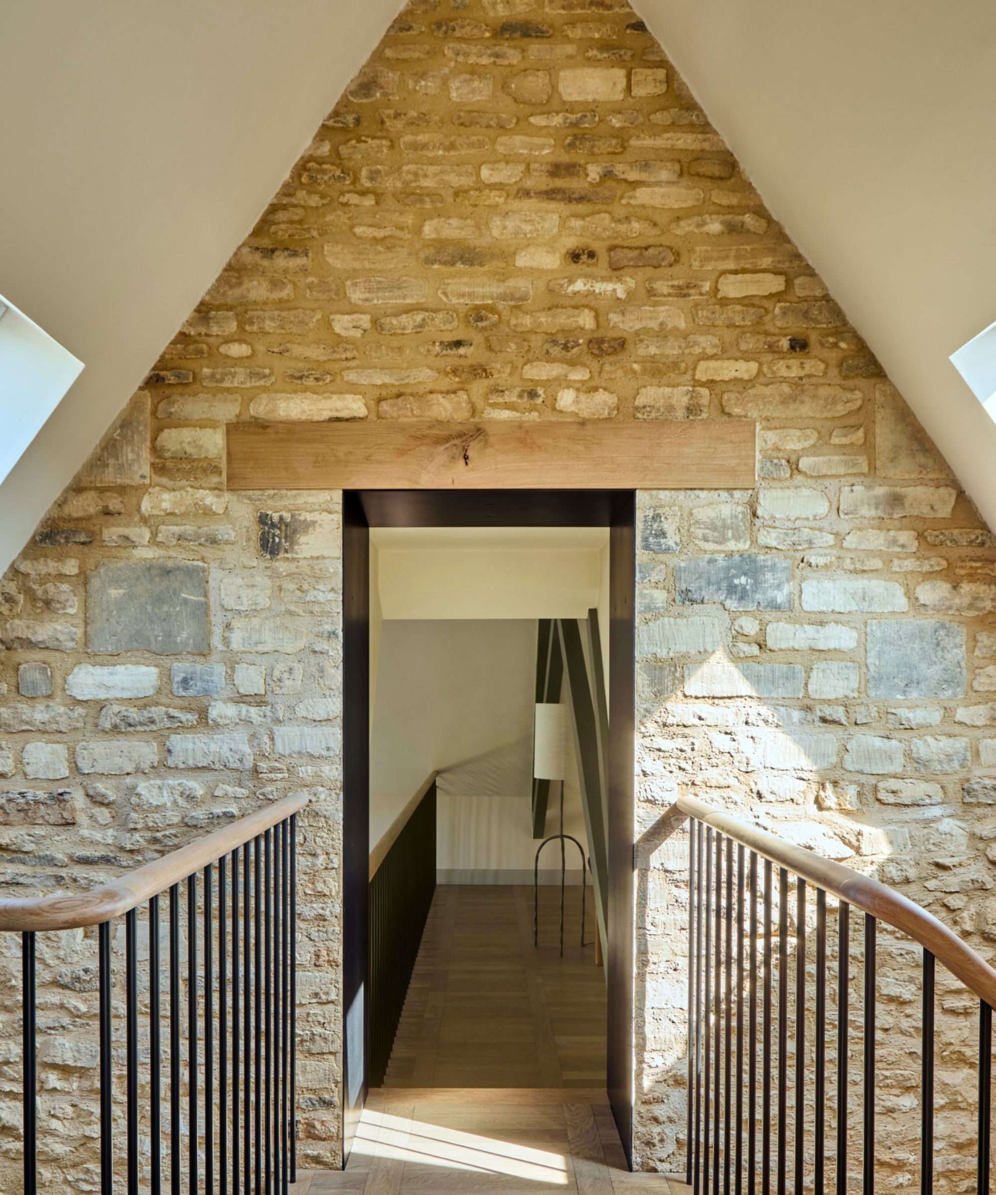 A landing inside a converted chapel with exposed Cotswold stone walls rising to a pitched apex, a black-framed doorway with oak lintel and wooden handrails.
