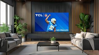 A TCL QD-Mini LED TV wall mounted in a room. Below it is a media cabinet, herringbone floor with a white rug, and a coffee table which is flanked by two sofas. The TV is showing an NFL player next to the NFL and TCL logos. QD-Mini LED is also written on the screen.