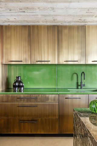 Image of a kitchen countertop and cabinetry. The ceiling is made of concrete and the cabinets are an aged brass material. The countertops and backsplash are made from a lime green prolave, lava stone material.