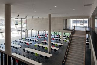 A large classroom with long desks, different colour chairs, large windows and a staircase going up on the right hand side.