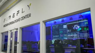 A control room with blue lights and lots of computer panels sits behind glass with a sign that reads Firefly Mission Operations Center above it.