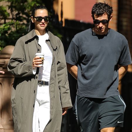 dua lipa wears a trench coat, white tee, belt, and white jeans while walking with callum turner.