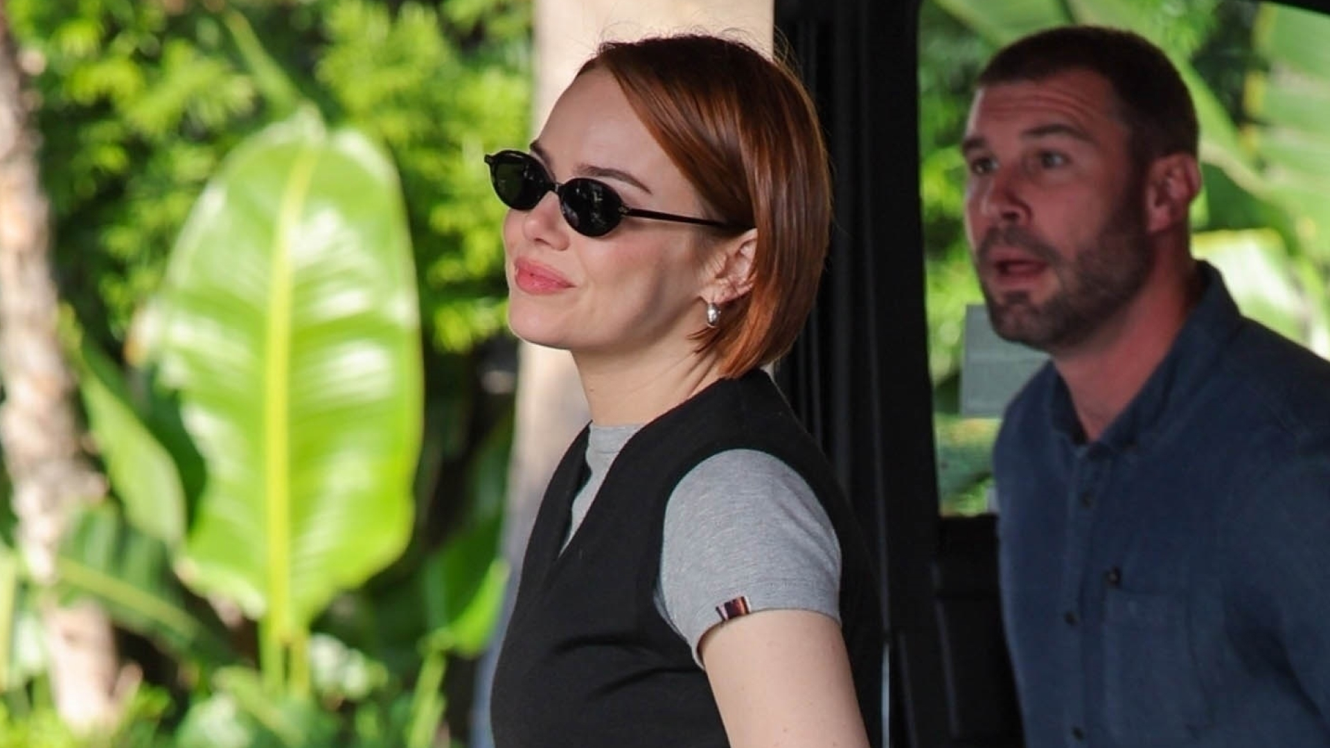Emma Stone wears black parachute pants, black strappy heels, a black preppy vest, a gray t-shirt, and carries a Louis Vuitton bag