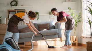 A man lifting a sofa so his friend can vacuum underneath