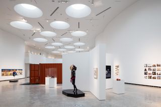 Exhibition viewing area, white room with marble floor, mannequin exhibit, white divider walls, metal orange container with door open, circular lights on the ceiling, artwork photographs on the right hand side wall