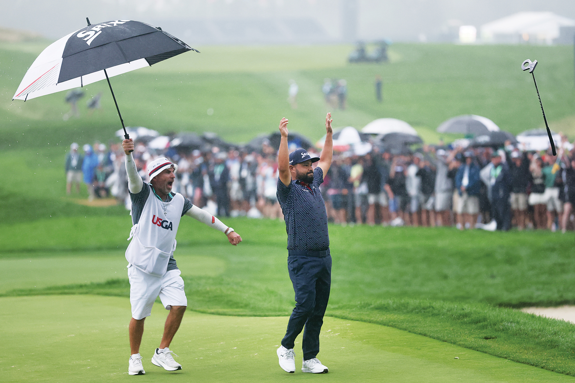 JJ Spaun throwing his putter in celebration after winning the US Open in 2025, with his caddie celebrating by raising his umbrella aloft in the rain