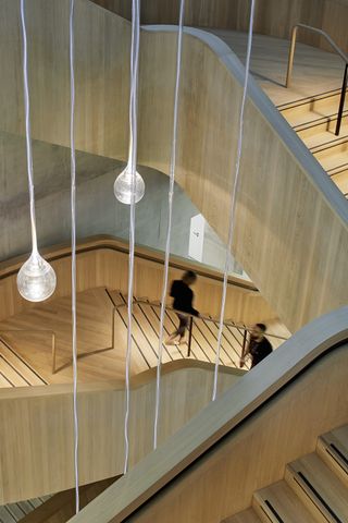Wood design staircase at AstraZeneca's Discovery Centre by Herzog de Meuron with glass ceiling lights hanging from the ceiling. Blurred image of a woman in all black going down the stairs and a man in all black coming up the stairs
