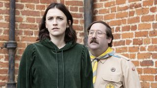 Charlotte Ritchie in a green top as Alison and Jim Howick in a scout master's outfit as Pat stand outside Button House in Ghosts.