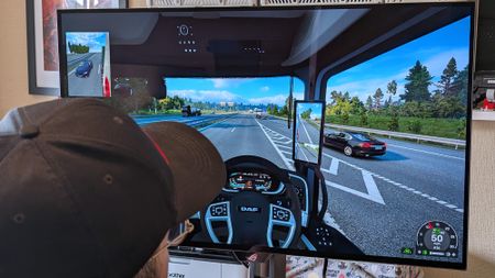 Windows Central's Ben Wilson wearing a black cap sits at a desk playing Euro Truck Simulator 2, with the ASUS ROG Strix OLED XG32UQWMS monitor showing a DAF truck interior.