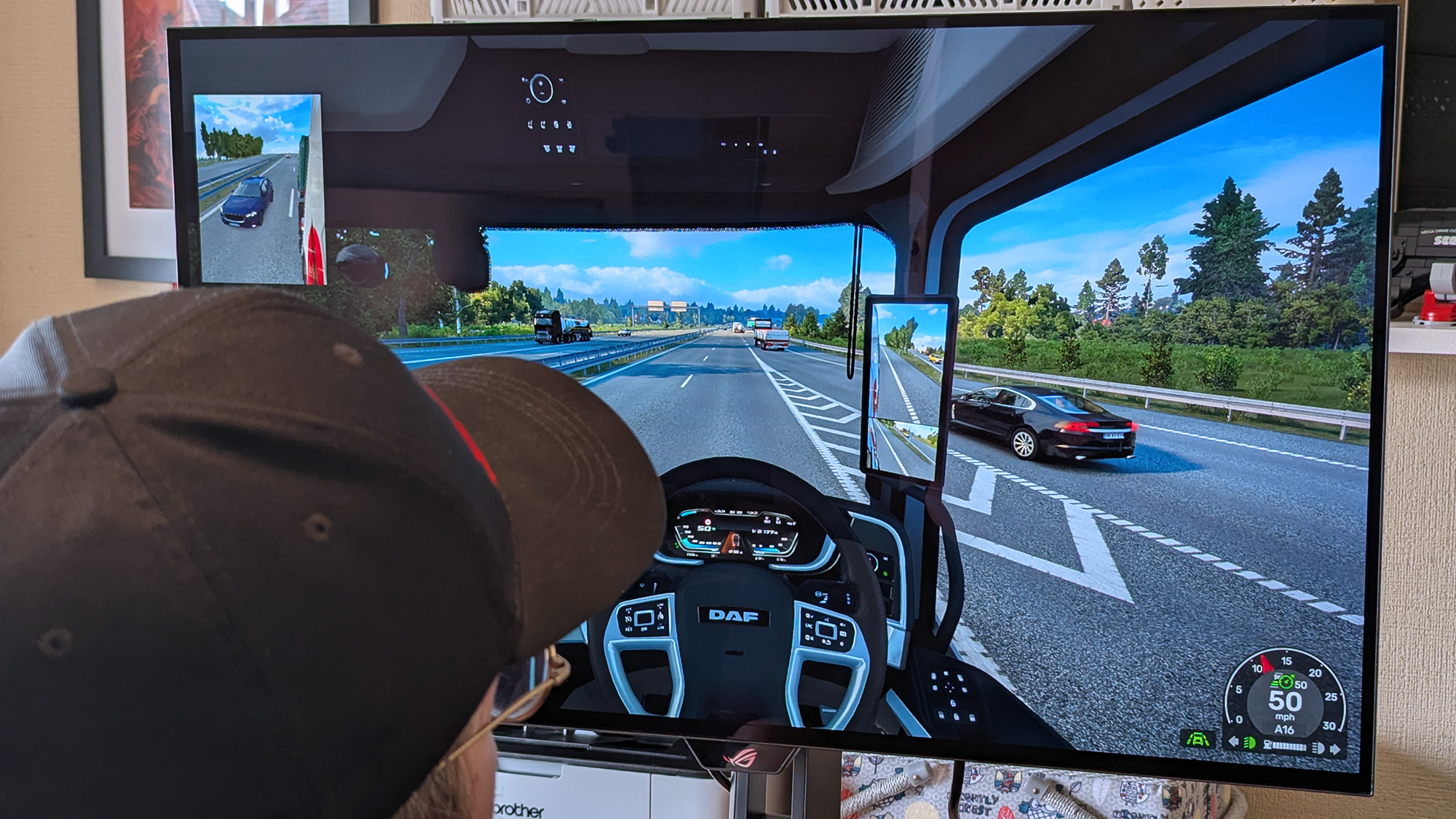 Windows Central's Ben Wilson wearing a black cap sits at a desk playing Euro Truck Simulator 2, with the ASUS ROG Strix OLED XG32UQWMS monitor showing a DAF truck interior.