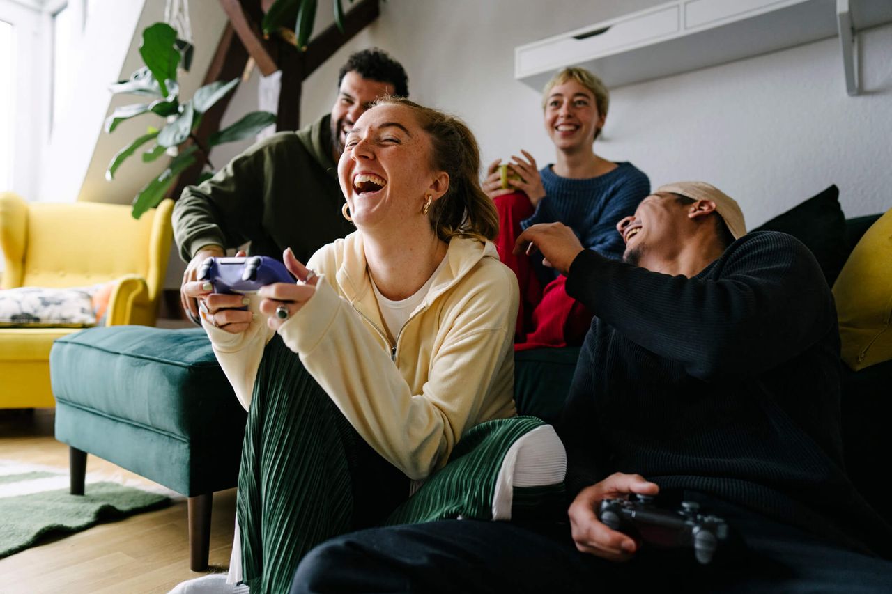 A group of four friends, two friends are sat on a couch and two are sat on the floor and they&#039;re playing video games together whilst laughing