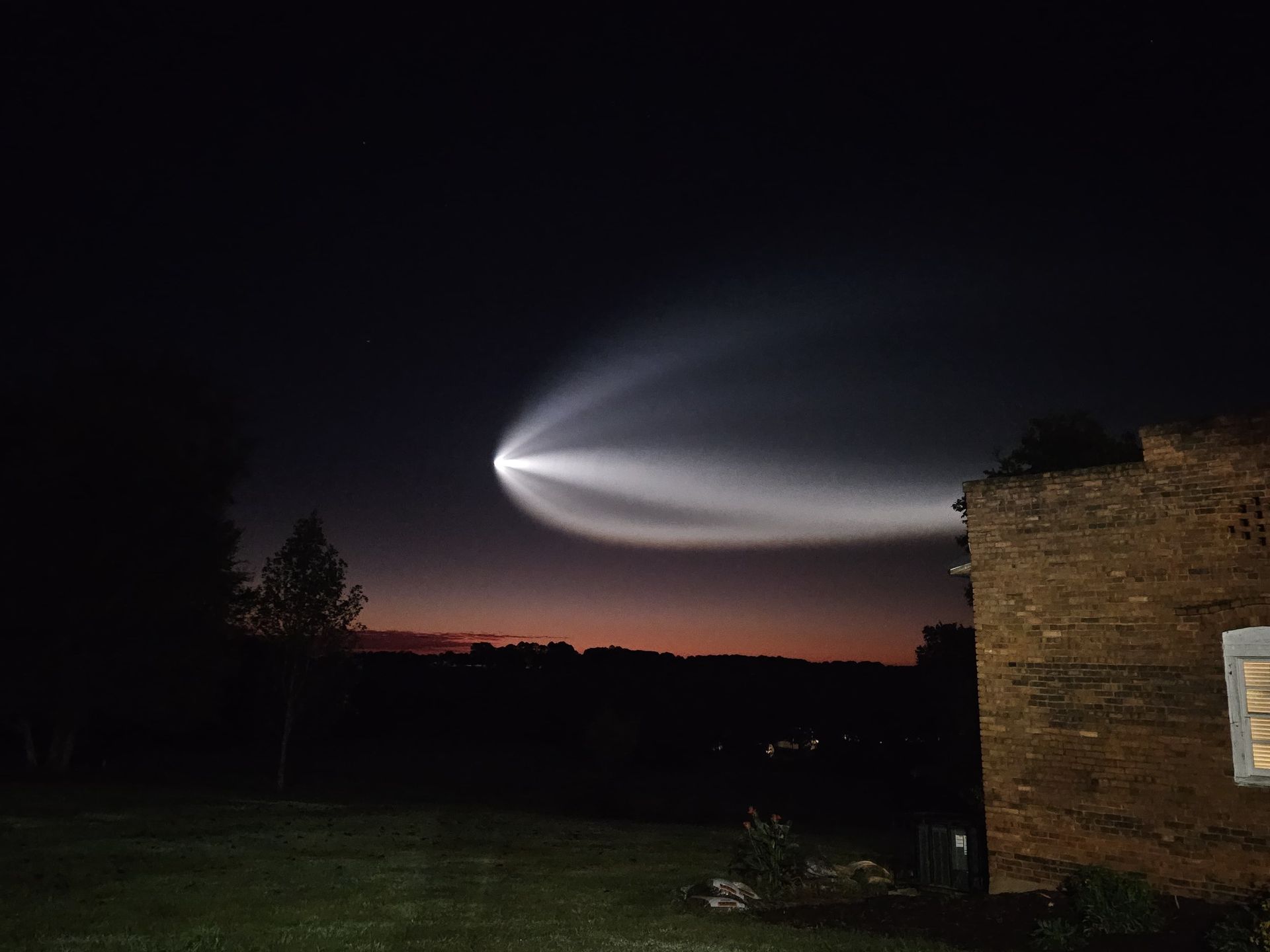 a white streak illuminated in the sky flies over a dark landscape and a brick house. 