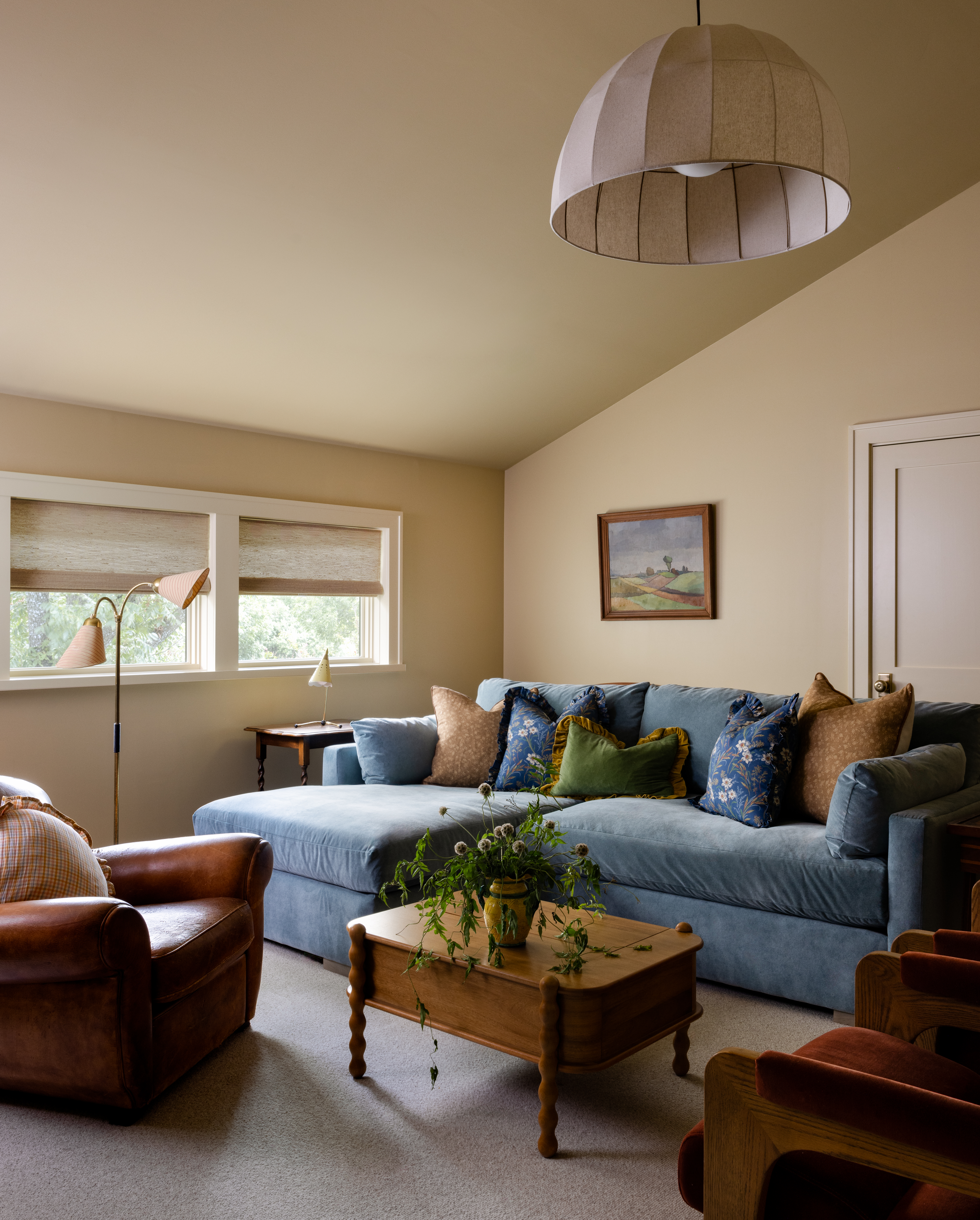 A photo of a cozy, sunlit living room with a large blue velvet sectional couch, a leather armchair, a wooden coffee table with a plant, and a large pleated dome lamp.