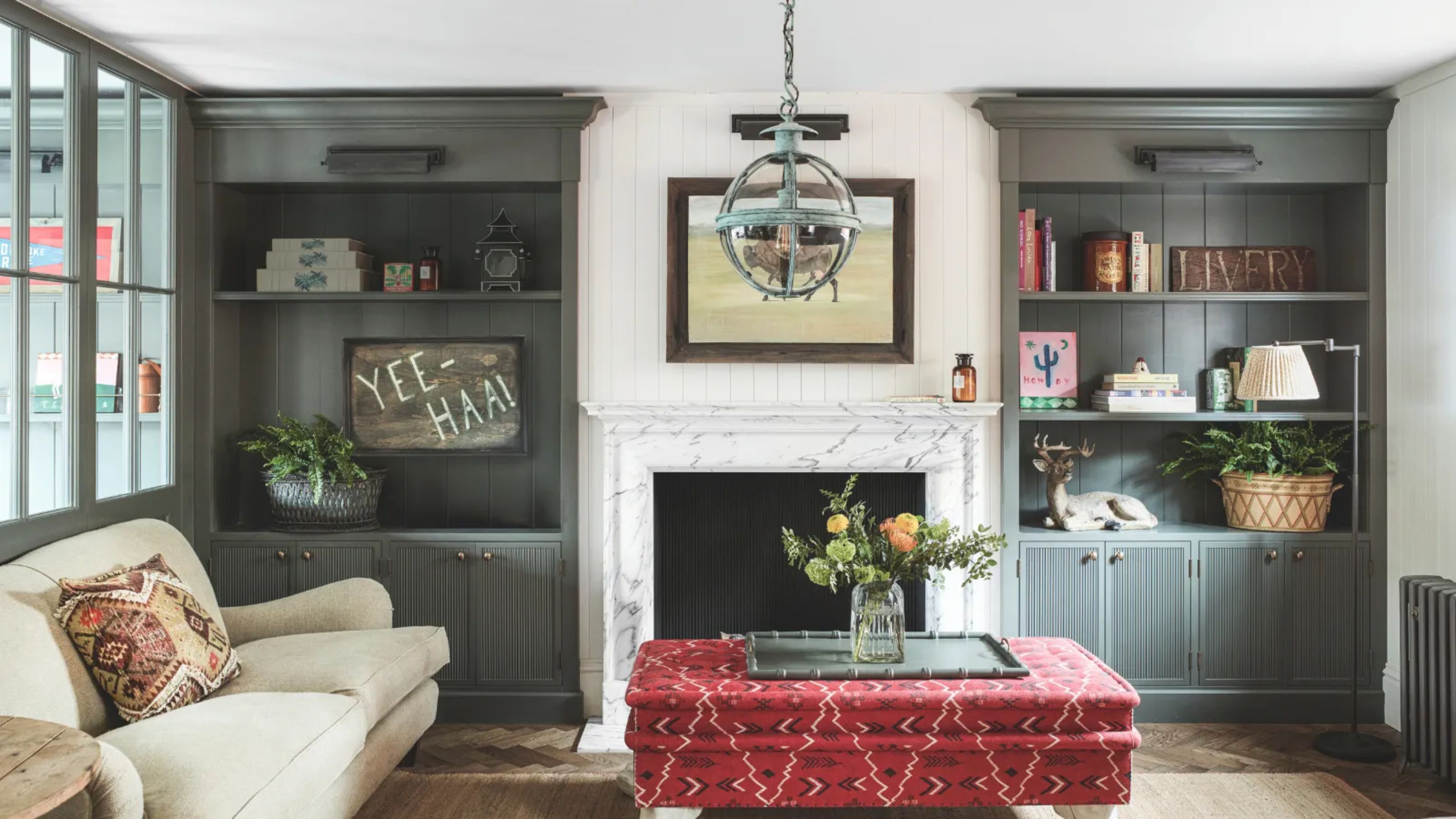 A rustic living room with dark gray built-in shelves, a red ottoman, and a tan couch