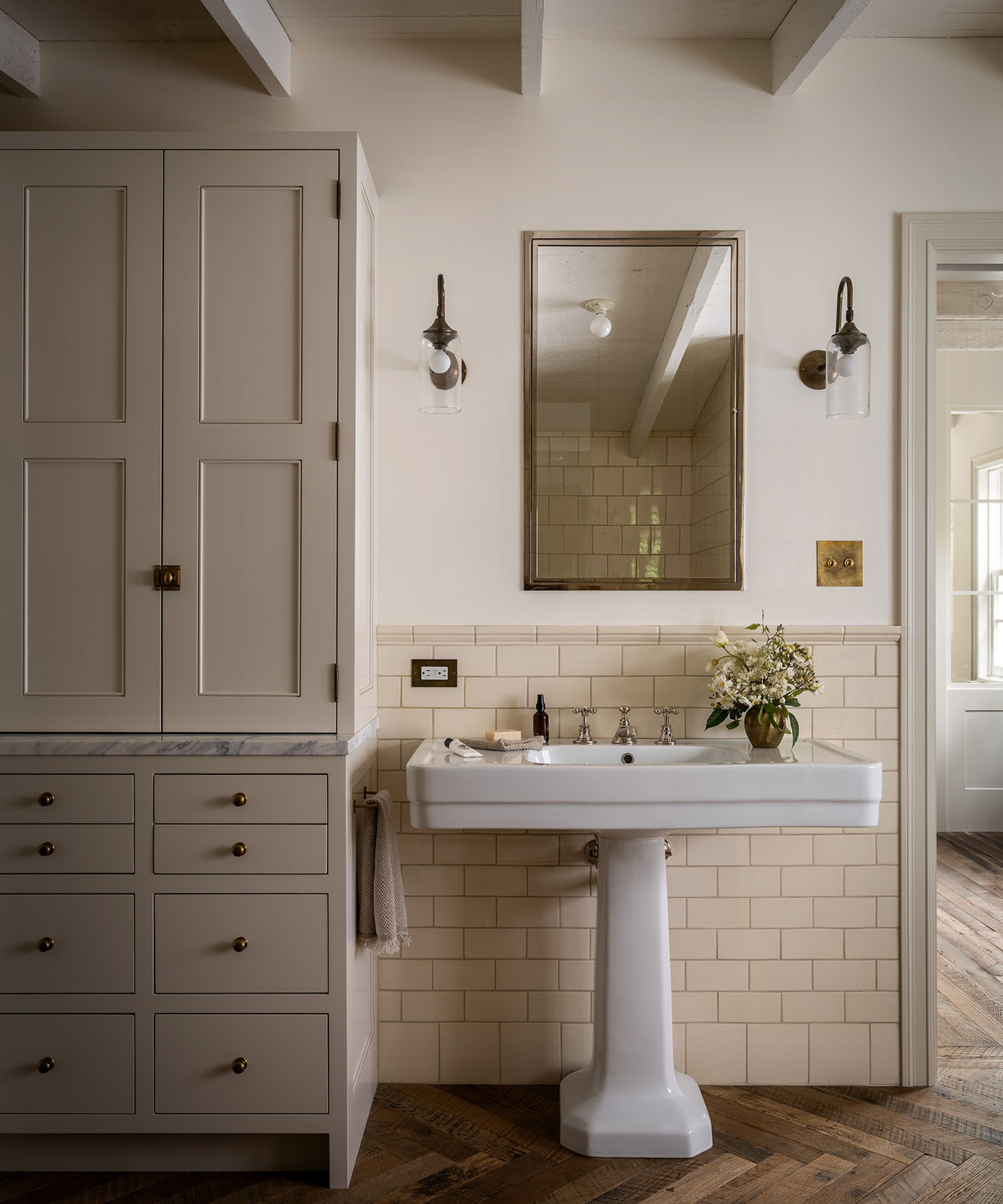 a warm white bathroom with herringbone wooden floor and rustic ceiling beams with a pedestal sink and custom cabinetry