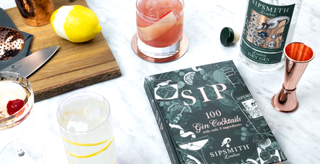 Cocktails, ingredients and cocktail-making tools scattered on a marble surface next to Sipsmith's SIP book