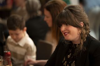 Tosh (Alison O'Donnell) at a party with various people out of focus in the background, including a young boy. There are cans empty glasses and cans on the table in front of her. She is smiling a little uncomfortably.