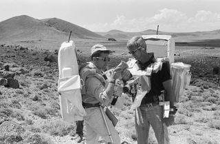 astronauts Eugene A. Cernan (right) and Harrison H. Schmitt (left) are wearing large white "backpacks" and inspect a rock sample.