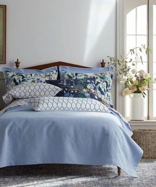 Blue and white bedding with floral shams on a bed against white walls; to the right, a large vase of flowers.