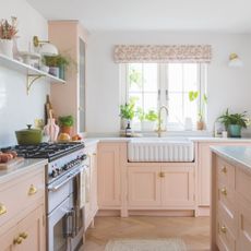 White painted kitchen with baby pink cabinets and a white ceramic sink underneath a window