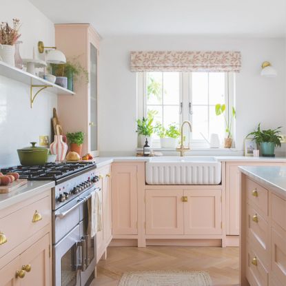 White painted kitchen with baby pink cabinets and a white ceramic sink underneath a window