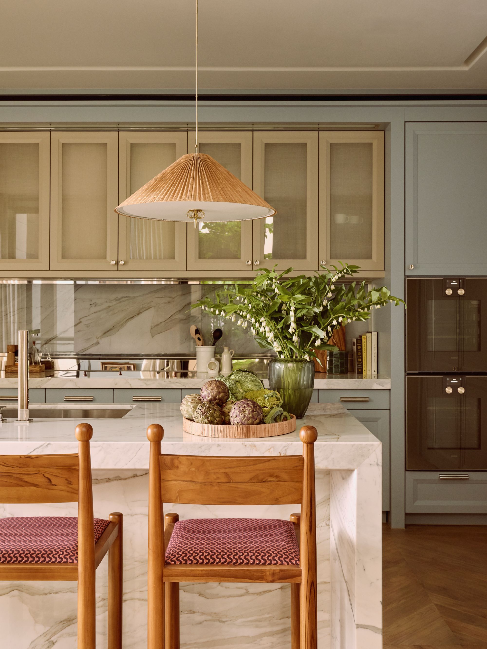 a kitchen with a white marble countertop, wooden barstools with red seat cushions, a decorative tray holding vegetables and a vase of flowers. Behind is a wall of cabinets painted in sage green and light blue