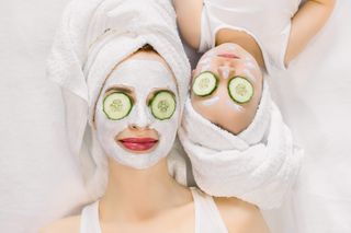 Mother and daughter in white bath towels on head and with slices of cucumber on their eyes. Woman has white facial mask on her skin.