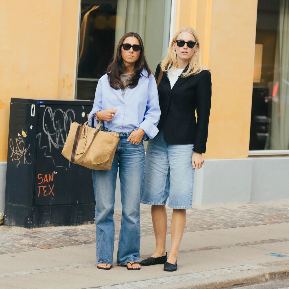 Two women pose in Copenhagen. One woman wears a button-down and jeans, and sandals. The other wears a black blazer and jorts with flats. 
