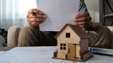 A model of a home sitting on top of paperwork with a man reviewing paperwork in the background.