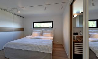 Interior view of a bedroom at Trekronekabin featuring white walls, a window, wood flooring, a bed with white and grid style pillows and linen, a white sliding wardrobe with wood panels, a mirror and white bar spotlights. The en-suite bathroom can also be partially seen