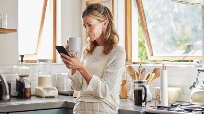 Woman standing in kitchen holding cup of coffee and phone in hand with appliances behind her, researching what causes constipation