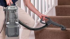 Man wearing beige slack pants operating a Shark lift-away floorcare vacuum cleaner on brown carpeted stairs