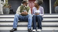 An older couple with coffee sit on the steps of their home and smile at each other.