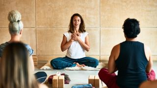 Woman sitting down at the front of yoga class as instructor, crossed legged and hands together, eyes closed