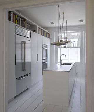 A white kitchen with pendant lighting above a narrow island, floor to ceiling cabinets and head-height book shelf