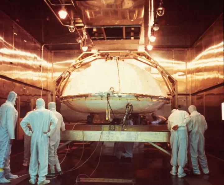 five people in white protective clothing surround a disk-shaped white spacecraft inside a clean room