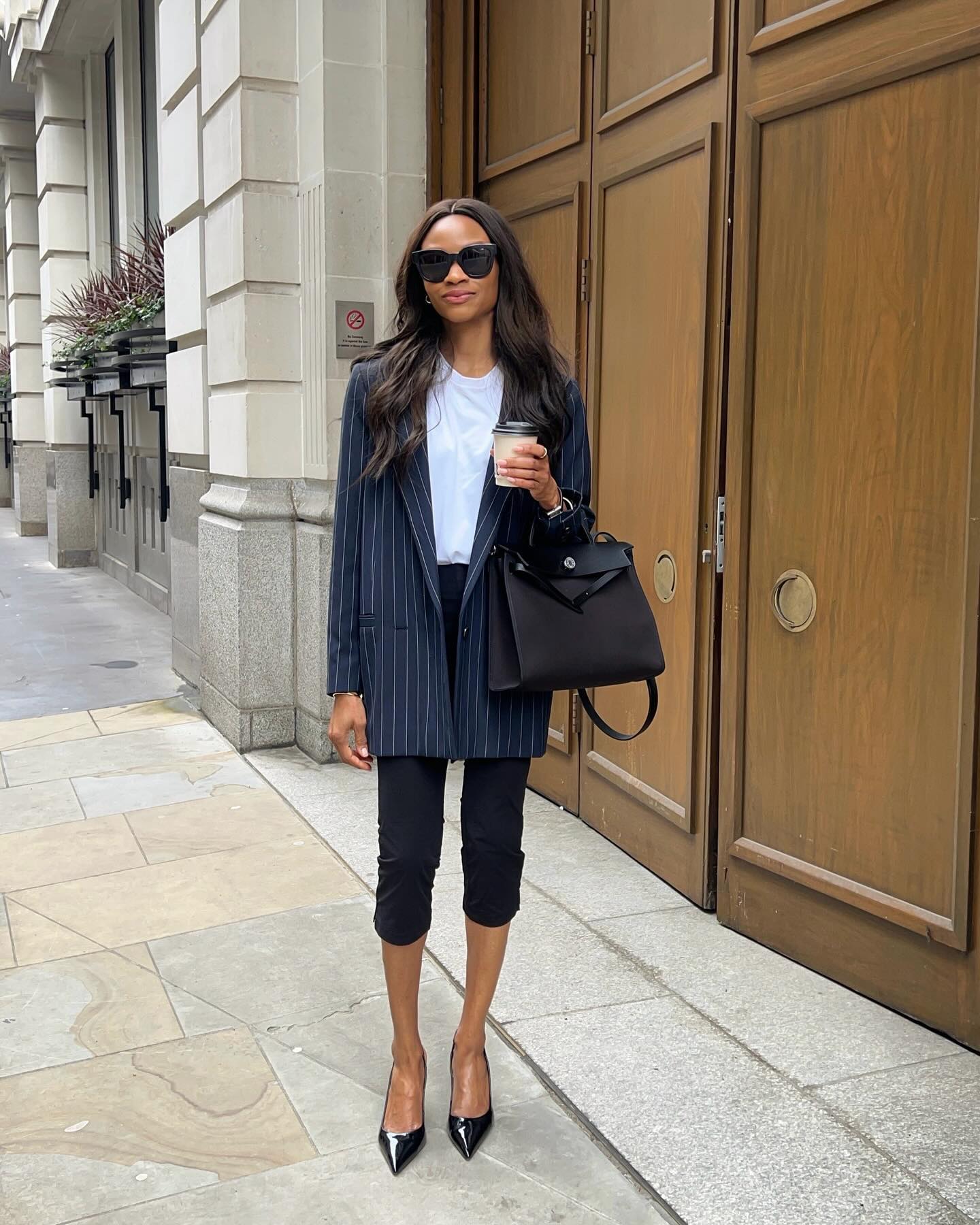 A woman wears a stripe blazer, white tee, black capri pants, and black pumps.