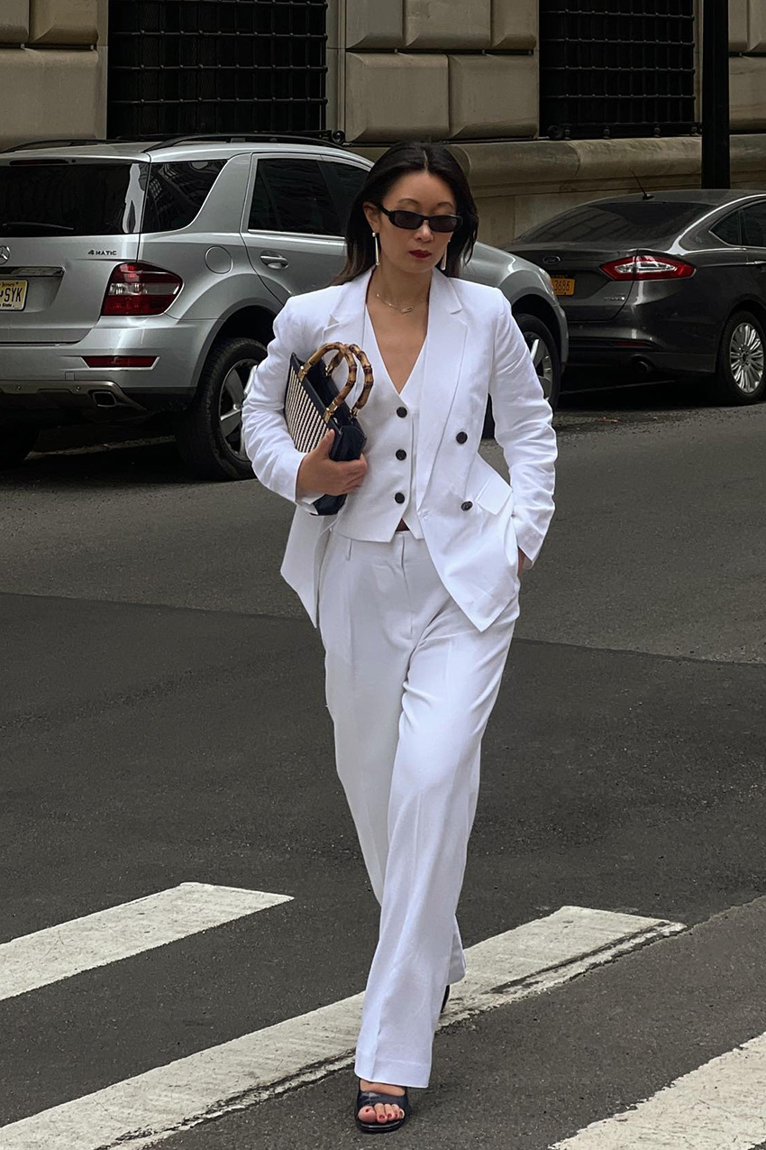 White outfit idea for summer is shown in a photo of a woman crossing the sidewalk in New York wearing black sunglasses with a three-piece white suit, a black Gucci bag, and black heeled mules