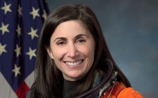 a woman in a NASA flight suit smiles beside an American flag