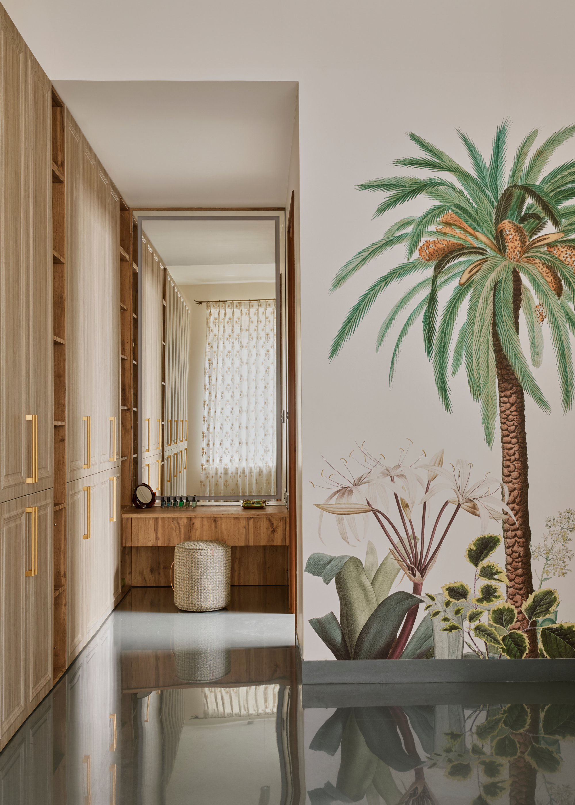 A bedroom with a botanical mural by a row of wood cupboards and a large vanity with a round pouff