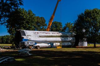 a large white rocket lays on its side surrounded by trees and a crane truck